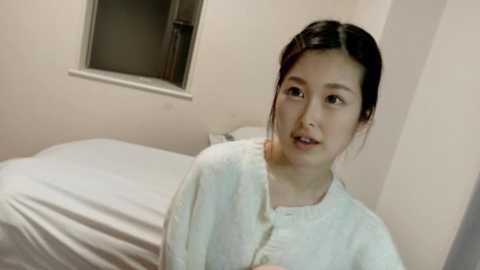 A video of a young Asian woman with dark hair tied back, wearing a white hospital gown, looking slightly distressed, in a sparsely furnished room with a single bed and a small window.