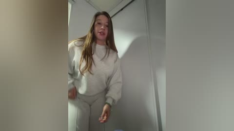 Video of a young woman with long brown hair, wearing a white knit sweater and matching pants, standing in a narrow, dimly lit hallway with white walls.