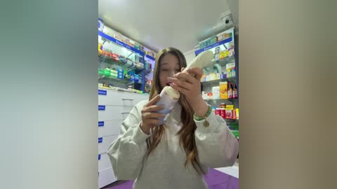Video of a young woman with long brown hair in a pharmacy, holding a bottle of milk, wearing a white hoodie and a green wristband. Shelves stocked with colorful medicines in the background.