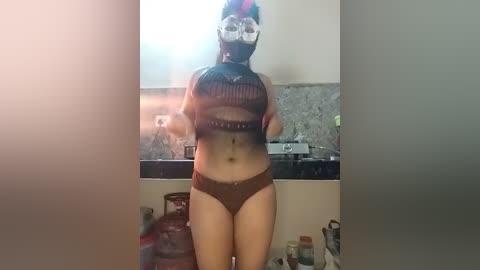 Video of a woman wearing a black, see-through mesh mask, brown lingerie, and high-waisted panties, standing in a bathroom with a sink, faucet, and metallic objects.