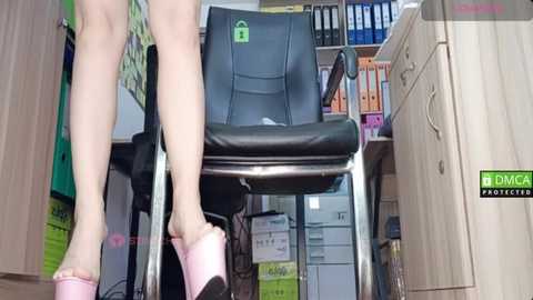 Video of a person's legs in pink high-heeled sandals, standing in front of a black office chair with a green lock symbol, surrounded by cluttered office shelves and cabinets.