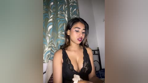 Video of a young woman with medium brown skin, dark hair, and wearing a black lace bra, seated indoors, holding a stuffed toy, surrounded by teal and gold patterned curtains and a metal-framed bed.