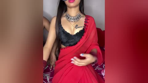 Video of an Indian woman in a red sari, black lace bralette, silver necklace, and maroon dupatta, with long dark hair, smiling, holding a phone.