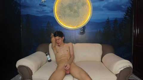 Video of a naked, fair-skinned man with a small penis, sitting on a beige couch in a dimly-lit room with a moon mural and a glowing moon lamp.