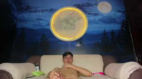 Video of a shirtless man with short dark hair lying on a beige couch, illuminated by a glowing moon-shaped lamp. Background features a dark blue night sky with stars and mountains.