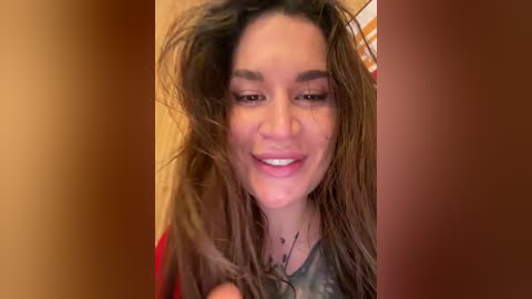 A video of a young woman with long, wavy brown hair, smiling, wearing a red top, against a warm-toned background.
