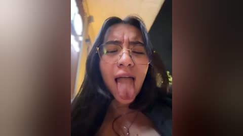 Video of a young woman with long dark hair, wearing glasses and a black top, sticking her tongue out, indoors with blurred background.