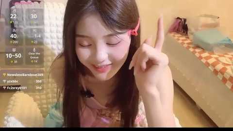 Video of a smiling East Asian woman with long black hair and a red hair clip, wearing a pink dress, making a peace sign, in a brightly lit bedroom.