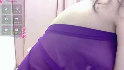 A video of a woman with fair skin and long black hair, wearing a sheer purple and yellow strapless dress, leaning against a pink wall with a fitness tracker display showing steps and calories.