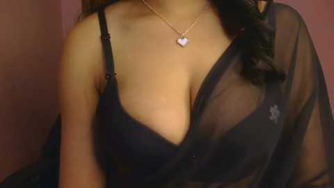 Video of a woman with medium brown skin, wearing a sheer black blouse that reveals a black bra, adorned with a delicate gold necklace.