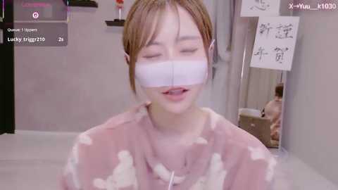 A video of a young Asian woman wearing VR goggles, pink top, and white tape over her mouth, in a sterile room with white walls and medical equipment.