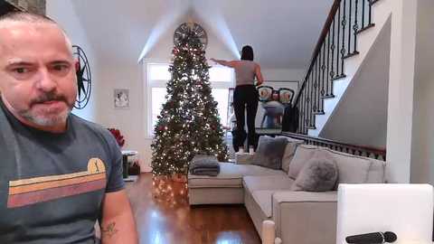 Media: Video of a man with a mustache in a gray T-shirt, a woman in a tank top and leggings, and a Christmas tree in a modern living room.