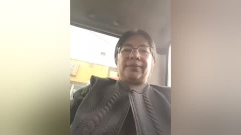 A video of a middle-aged Asian man with glasses, wearing a black quilted jacket, smiling in a dimly lit room with a yellow wall and a window behind him.