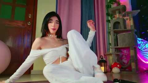 Video of a young Asian woman with black hair, wearing off-shoulder white outfit, sitting on a floor, surrounded by colorful curtains, a cat tree, and lanterns.