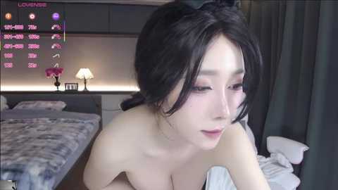 A video of an East Asian woman with long black hair, wearing makeup, in a bedroom. She's topless, with a pink flower on the bed, a digital display in the background.