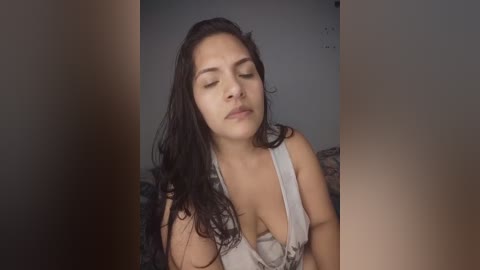 Video of a young Latina woman with long dark hair, medium skin tone, wearing a low-cut, white lace camisole, eyes closed, looking serene. Background is a plain, dimly lit room.