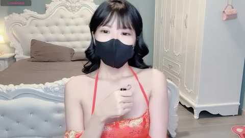 Video of a young Asian woman with fair skin and long black hair, wearing a red lace dress and black face mask, sitting on a bed in a pastel-colored bedroom.