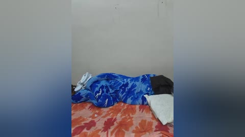 Video of a sleeping person, covered with a blue and white patterned blanket, on a bed with orange and white floral sheets. The background is a plain, off-white wall.
