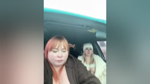 Video of two women in a car; one with red hair, wearing a dark jacket, and the other with blonde hair and a white jacket.