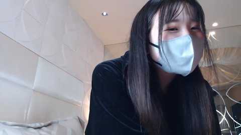 A video of an East Asian woman with long black hair, wearing a surgical mask and black clothing, leaning forward in a modern, beige-tiled room.