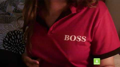 Video of a woman with medium skin tone wearing a red polo shirt with the word \"BOSS\" on the chest. Background includes a bed with a black and white striped comforter and a fan.