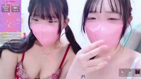 A video of two young East Asian women with light skin and dark hair, wearing matching red lace bras, blowing pink bubble gum bubbles.