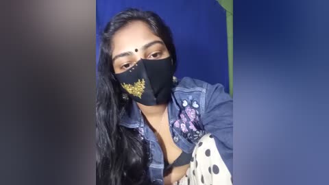 Video of a South Asian woman with long black hair, wearing a black face mask, denim jacket with floral embroidery, and a black top, against a blue background.