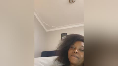 Video of a smiling Black woman with a large afro, lying on a bed with white sheets and a dark headboard, in a dimly lit room with a small framed picture on the wall.