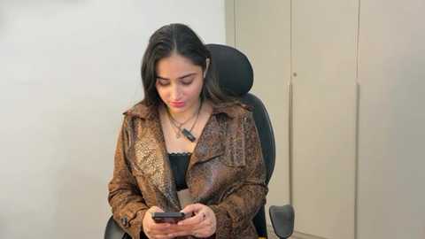 Media: Video of a young woman with medium skin tone and dark hair, wearing a brown leather jacket over a black top, sitting in a black office chair, intently looking at a smartphone.