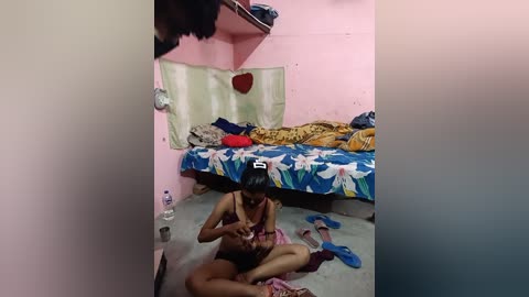 Video of a small, cluttered room with pink walls. A woman with dark skin and medium build sits cross-legged on the floor, feeding a baby. A bed with floral sheets and a red pillow is in the background.