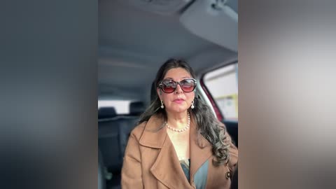 Video of a woman in a car, wearing large sunglasses, pearl necklace, and a tan coat, with wavy brown hair, seated in a dark interior, slightly out of focus.