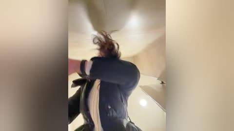 A blurry video of a person with shoulder-length brown hair, wearing a black jacket and black pants, running away from the camera in an indoor setting with beige walls and a white ceiling.