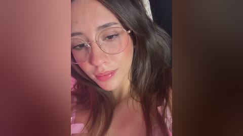 Video of a young woman with light skin, brown hair, and glasses, smiling gently. She wears a pink shirt, partially visible, and the background is blurred.