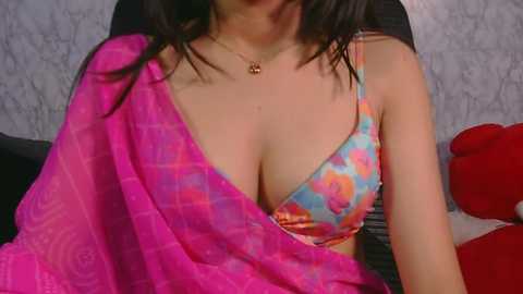 Video of a woman with medium skin tone and dark hair, wearing a colorful floral bra and a pink shawl, sitting on a black chair against a white, patterned wall.