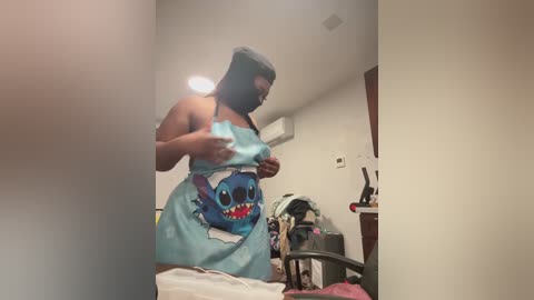 Video of a plus-sized Black woman with dark skin and long hair, wearing a blue Stitch-themed apron, standing in a cluttered bedroom with a bed, a desk, and a white wall.