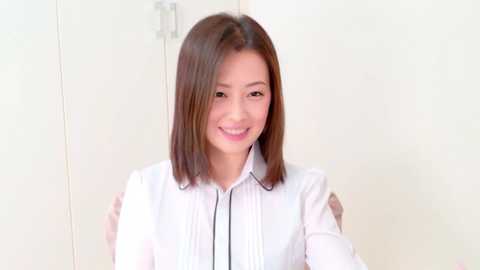 Media: Video of an East Asian woman with straight, shoulder-length brown hair, wearing a white, button-down shirt with a black trim. She has a warm smile and stands in front of a plain, off-white wall.
