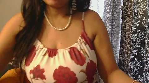 Media: Video of a woman with medium brown skin, wearing a red and white floral-patterned spaghetti strap top, pearl necklace, and long black hair, standing in a room with black and white floral-patterned curtains.
