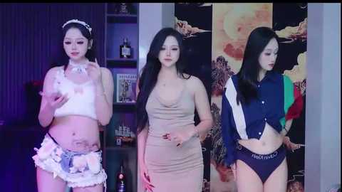Video of three East Asian women in provocative outfits: white lingerie, beige dress, and navy jacket with boxer shorts. Background features a dark room with shelves and wall art.
