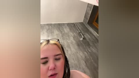 Video of a young woman with blonde hair, wearing glasses, lying on a wooden floor with a metallic object beside her. The room has white walls and a brown wooden door.