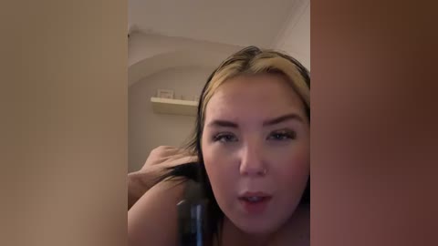 Video of a young woman with light skin and blonde-streaked hair, seen from a slight angle. She's in a bathroom, holding her hair, and looking directly at the camera.