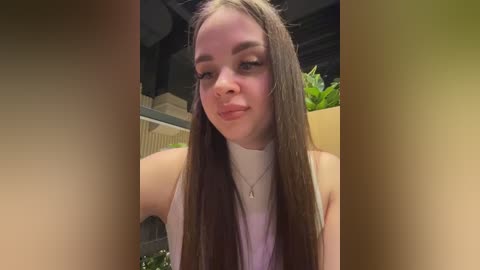Video of a young Caucasian woman with long, straight brown hair, wearing a sleeveless white top, standing in a modern, indoor setting with plants in the background.