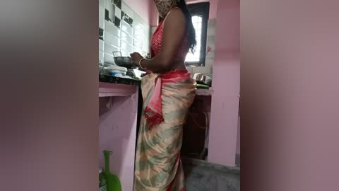 Video of a dark-skinned woman with braided hair in a red and gold sari, standing in a pink-tiled kitchen with a green broom.