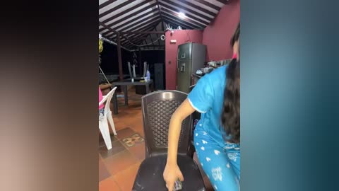 Video of a rustic outdoor space with a child in a blue shirt playing table tennis on a black table, surrounded by colorful plastic chairs and a red wall, under a thatched roof.