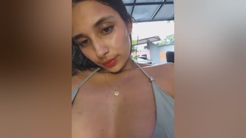 A close-up video of a young woman with olive skin, dark hair, and full lips. She wears a low-cut, light blue top, showing cleavage. The background is blurry, showing a patio area with a green plant and a building.