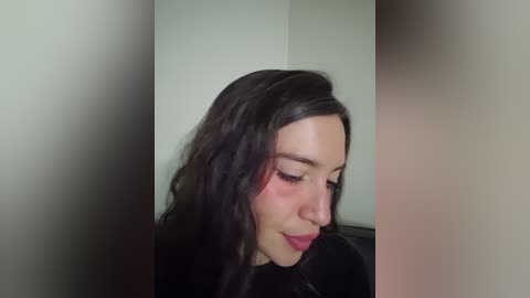 Video of a young woman with long, wavy dark hair, fair skin, and closed eyes, wearing a black top, standing against a plain, off-white wall.