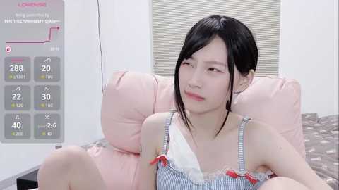 A video of an Asian woman with straight black hair, wearing a blue-and-white striped camisole, sitting on a pink couch, with a digital health monitor in the background.