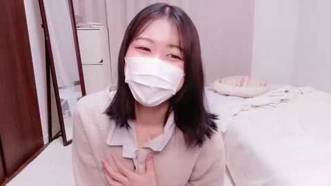 A video of an Asian woman with straight black hair, wearing a white mask, light jacket, and a white shirt, kneeling on a white floor in a minimalistic bedroom with a bed and white walls.