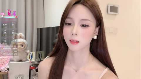 Video of a pale-skinned, slender East Asian woman with long brown hair, wearing a white spaghetti-strap top, sitting in a modern living room with a TV and gray curtains.