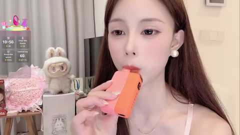 Media: A video of a young East Asian woman with long brown hair, wearing a pink camisole, eating an orange popsicle. Background features a pink bunny plush toy, a gift box, and a digital screen with game info.