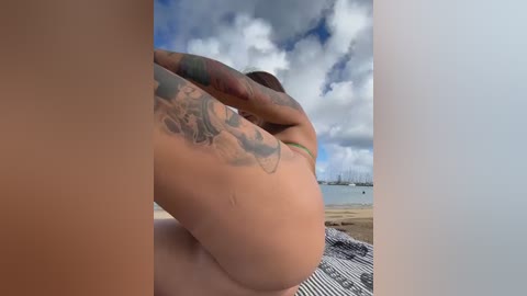 A video captures a nude woman with tan skin, multiple tattoos, and a green bikini top, sitting on a striped towel by the ocean under a cloudy sky.